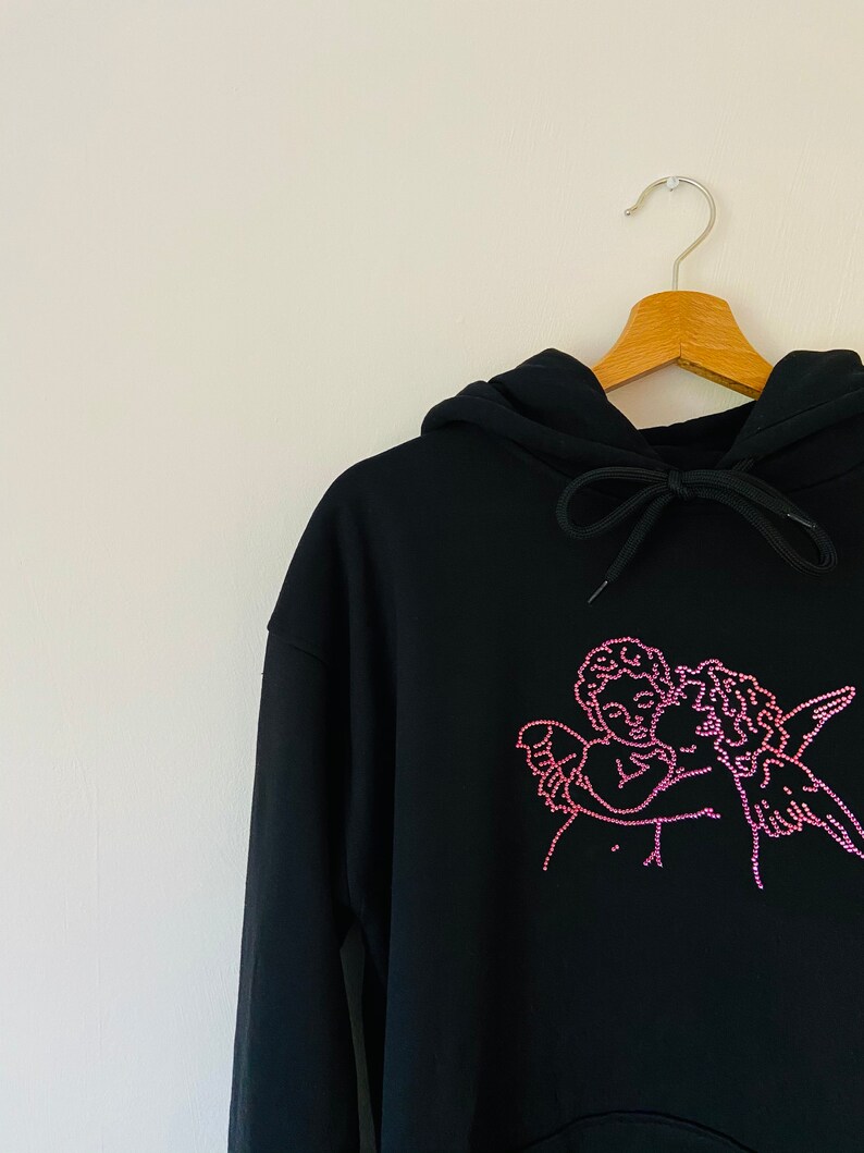 Pu&ograve; includere: Felpa con cappuccio nera con un design di due angeli che si abbracciano in strass rosa.