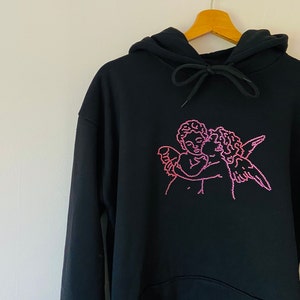 Pu&ograve; includere: Felpa nera con un design di due angeli che si abbracciano in strass rosa.