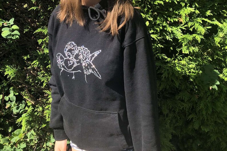 Pu&ograve; includere: Felpa nera con un design di due angeli che si abbracciano in strass bianchi.