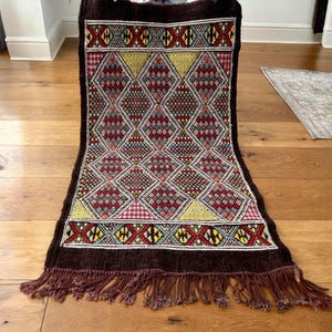 May include: A rectangular, hand-woven rug with a dark brown border and fringe. The central design features a repeating diamond pattern in red, yellow, and blue. The rug is placed on a wooden floor.
