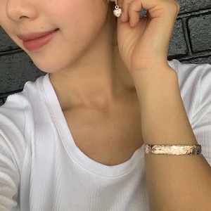 May include: A white shirt with a gold bracelet with a floral design. The bracelet is on the left wrist of the person wearing the shirt.