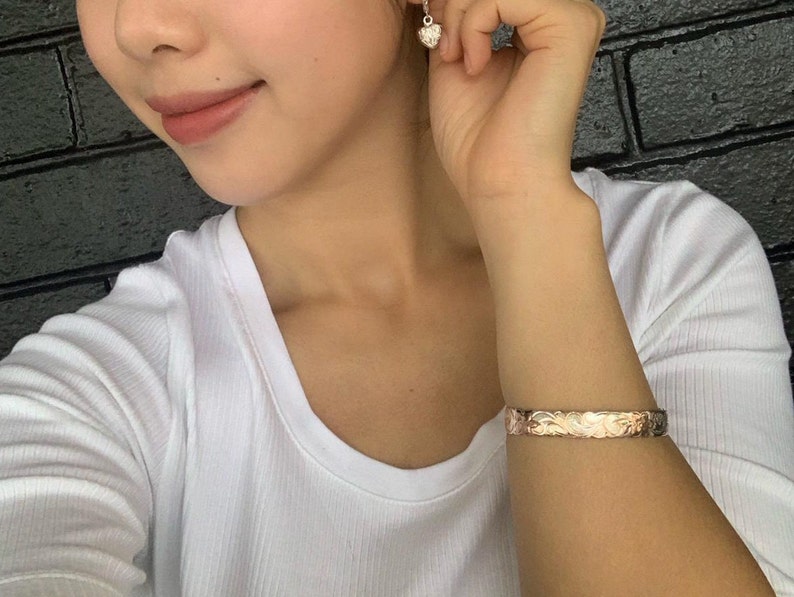 May include: A white shirt with a gold bracelet with a floral design. The bracelet is on the left wrist of the person wearing the shirt.