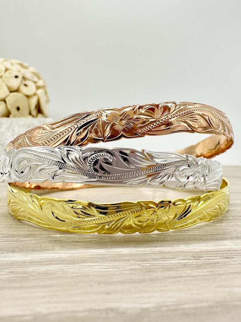 May include: Three Hawaiian flower lei bracelets in gold, silver, and rose gold. The bracelets are stacked on top of each other.