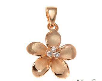Puede incluir: Un colgante de flor de plumeria en tono dorado con tres piedras de circonita cúbica transparentes en el centro. El colgante tiene un acabado cepillado.