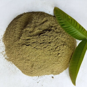 May include: A pile of green powder on a white surface with two green leaves beside it.