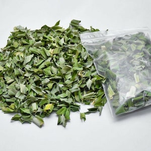 May include: A pile of dried, chopped green herbs next to a clear plastic bag filled with the same herbs. The herbs are a light green color and appear to be dried chives or a similar culinary herb. The background is plain white.