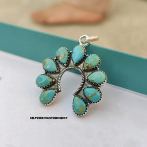 Pu&ograve; includere: Un ciondolo in argento con un design a ferro di cavallo, adornato con pietre turchesi a forma di goccia. Il ciondolo ha una cornice in argento dettagliata e strutturata e un piccolo anello per una catena. Il testo "SILVERRINGSTORESHOP" &egrave; visibile in basso.