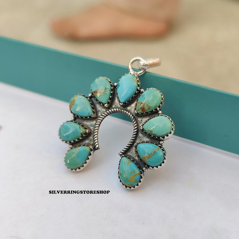 Pu&ograve; includere: Un ciondolo in argento con un design a ferro di cavallo, adornato con pietre turchesi a forma di goccia. Il ciondolo ha una cornice in argento dettagliata e strutturata e un piccolo anello per una catena. Il testo "SILVERRINGSTORESHOP" &egrave; visibile in basso.