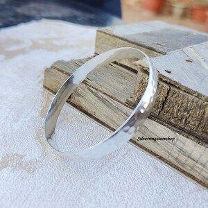 May include: A silver hammered bangle bracelet. The bracelet is smooth and shiny, with a hammered texture. The bracelet is lying on a wooden surface.