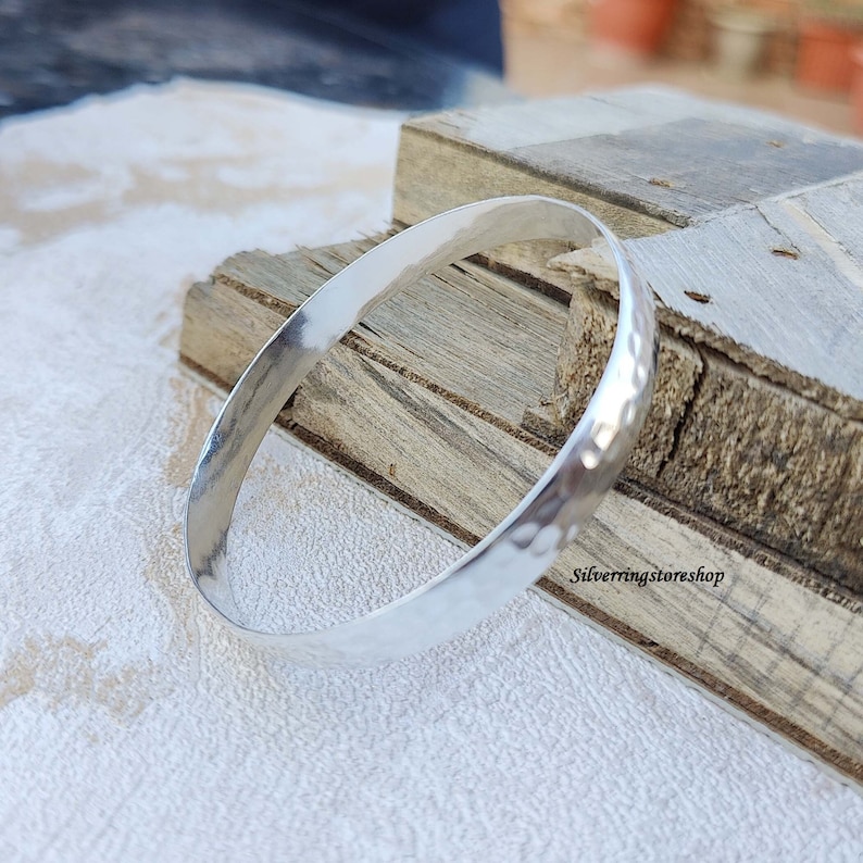 May include: A silver hammered bangle bracelet. The bracelet is smooth and shiny, with a hammered texture. The bracelet is lying on a wooden surface.