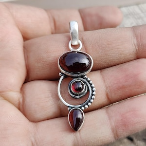 May include: A silver pendant with a red garnet gemstone. The pendant features a circular design with a smaller garnet in the center. The pendant is on a silver chain.
