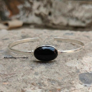 K&ouml;nnte beinhalten: Ein silberner Armreif mit einem glatten, polierten ovalen schwarzen Stein in der Mitte. Das Armband ist d&uuml;nn und offen gestaltet und hat eine metallische Oberfl&auml;che. Der Text silverringstoreshop ist auf der linken Seite zu sehen.