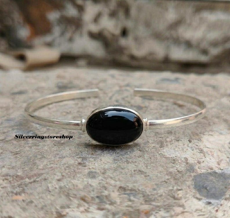 K&ouml;nnte beinhalten: Ein silberner Armreif mit einem glatten, polierten ovalen schwarzen Stein in der Mitte. Das Armband ist d&uuml;nn und offen gestaltet und hat eine metallische Oberfl&auml;che. Der Text silverringstoreshop ist auf der linken Seite zu sehen.
