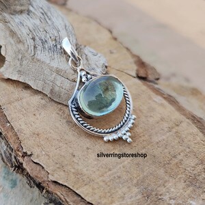 May include: A silver pendant with a light blue oval gemstone set in a detailed silver frame. The pendant has a bail for attaching to a chain.