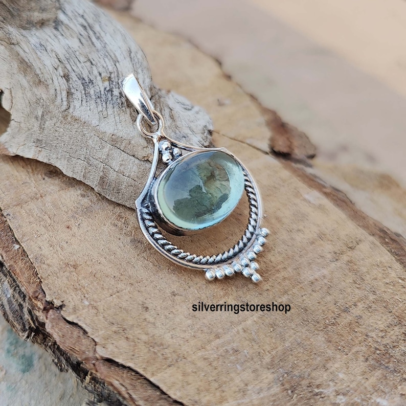 May include: A silver pendant with a light blue oval gemstone set in a detailed silver frame. The pendant has a bail for attaching to a chain.