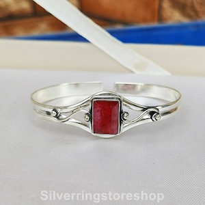 May include: A silver bangle bracelet featuring a red gemstone set in a rectangular frame. The bracelet has a delicate, ornate design with swirls and small beads.