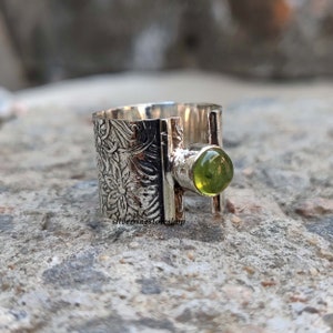 May include: A silver ring with a green gemstone set in a textured band. The band is decorated with a floral pattern.