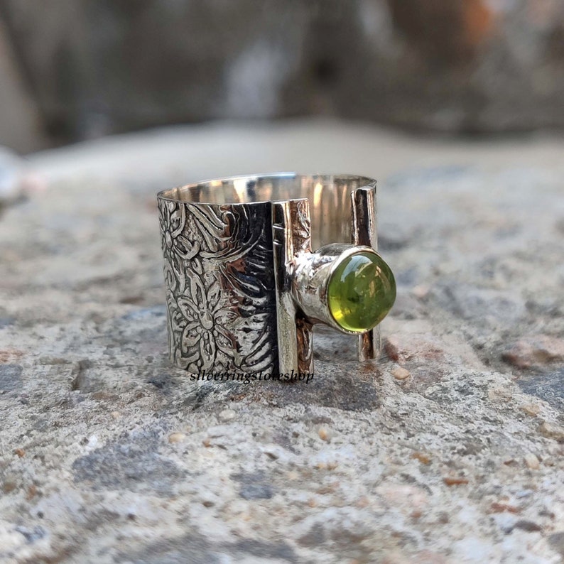 May include: A silver ring with a green gemstone set in a textured band. The band is decorated with a floral pattern.