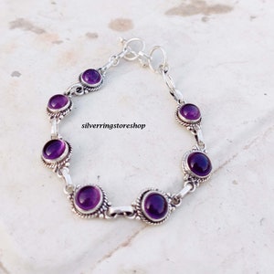 May include: A silver bracelet featuring seven round, deep purple amethyst stones. The stones are set in ornate, detailed bezels and connected by silver links. The bracelet is displayed on a light-colored surface, with the text "silverringstore shop" visible.