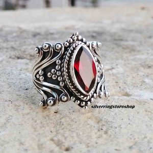 Puede incluir: Un anillo de plata con una piedra preciosa roja engastada en un dise&ntilde;o detallado y ornamentado. El anillo tiene una banda ancha con intrincados trabajos de desplazamiento y abalorios.