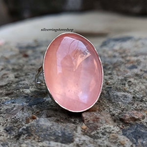 Puede incluir: Un anillo de plata con una gran piedra preciosa rosa de forma ovalada. El anillo está colocado sobre una roca gris y rugosa.