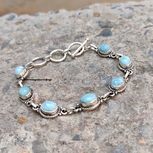 May include: A silver bracelet featuring oval, light blue gemstones. The bracelet has a toggle clasp and is composed of interconnected links. The gemstones are set in ornate, silver bezels, creating a delicate and elegant design.