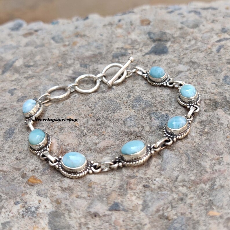 May include: A silver bracelet featuring oval, light blue gemstones. The bracelet has a toggle clasp and is composed of interconnected links. The gemstones are set in ornate, silver bezels, creating a delicate and elegant design.