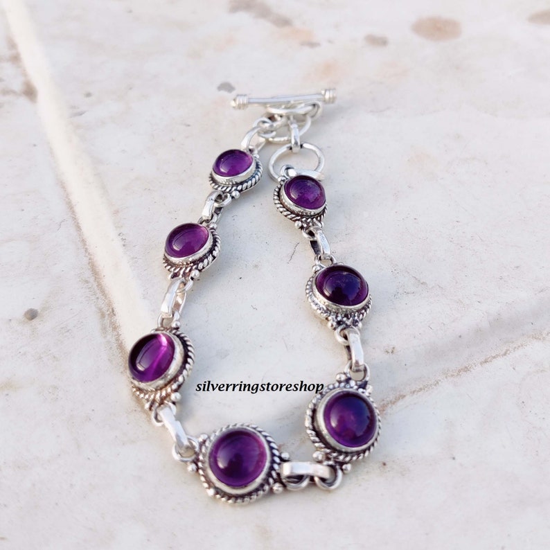 May include: A silver bracelet featuring seven round, deep purple amethyst stones. The stones are set in ornate, detailed bezels and connected by silver links. The bracelet has a toggle clasp. The text "silverringstore shop" is visible.