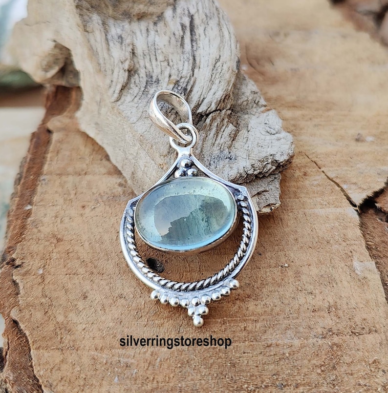 May include: A silver pendant with a light blue oval gemstone set in a decorative silver frame. The pendant has a bail for attaching to a chain.