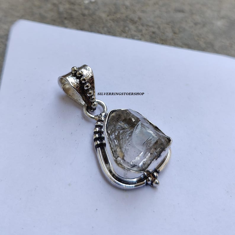 May include: A silver pendant featuring a clear quartz crystal set in a silver frame with beaded accents. The pendant is on a white background.  "SILVERRINGSTOERSHOP" is printed on the background.