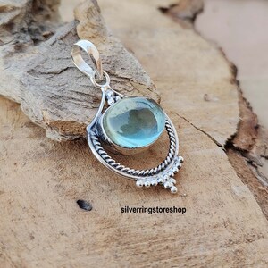 May include: A silver pendant with a light blue gemstone set in a decorative silver frame. The pendant has a bail for attaching to a chain.