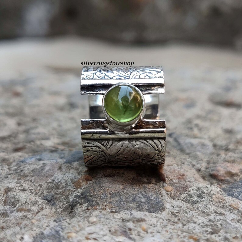 May include: A silver ring with a green gemstone set in a textured band. The band has a floral design and a silver stripe.