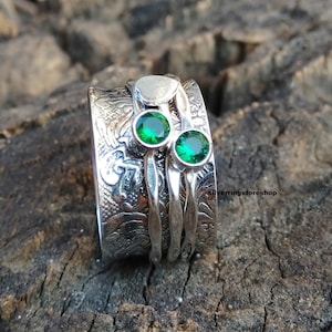 Może przedstawiać: Srebrny pierścień obrotowy z fakturowanym wzorem i dwoma zielonymi kamieniami szlachetnymi. Pierścień leży na drewnianej powierzchni.