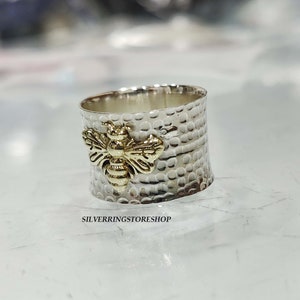 May include: A silver ring with a textured band and a gold bee design. The ring is a wide band with a hammered finish.
