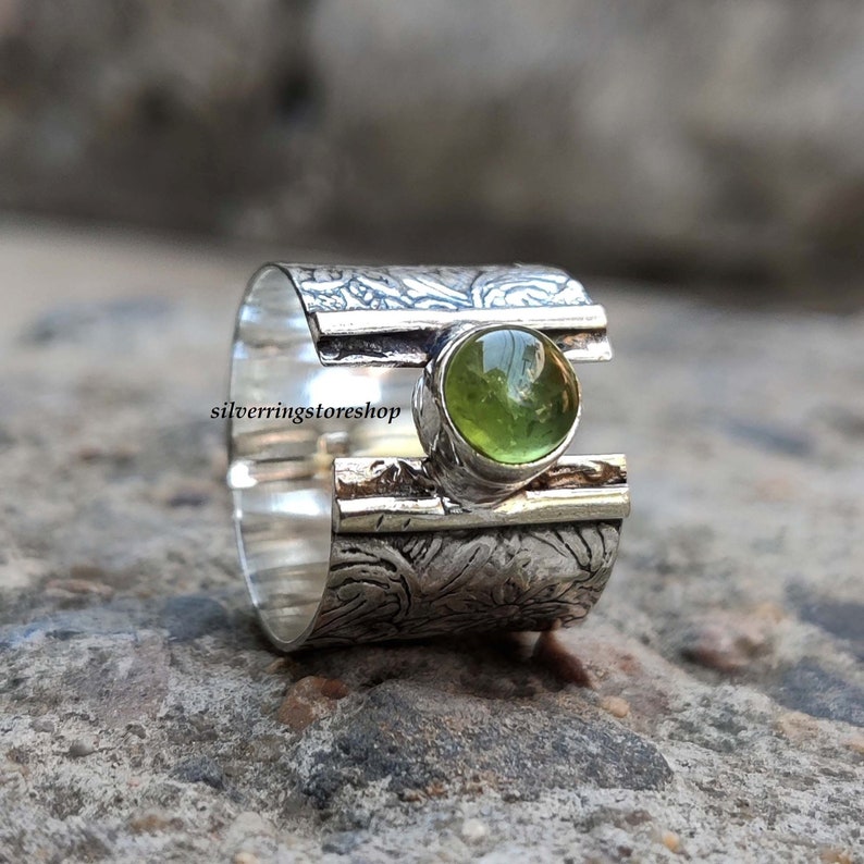 May include: A silver ring with a textured band and a green gemstone set in the center. The band is decorated with a floral pattern.