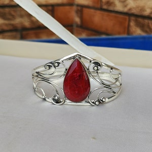 Puede incluir: Un brazalete de plata con una piedra preciosa roja en el centro. El brazalete tiene un diseño delicado y ornamentado.