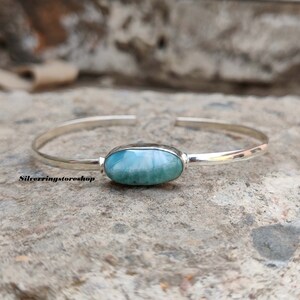 K&ouml;nnte beinhalten: Ein silbernes Armband mit einem zentralen, ovalen, blau-wei&szlig;en Larimar-Edelstein. Das polierte Metallband ist auf der R&uuml;ckseite offen. Der Text "Silverringstoreshop" ist in Schwarz auf der linken Seite des Armbands aufgedruckt.