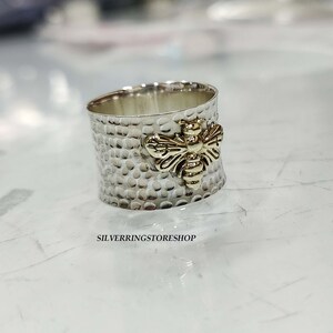 May include: A silver ring with a textured finish and a gold bee design. The ring is wide and has a hammered look.