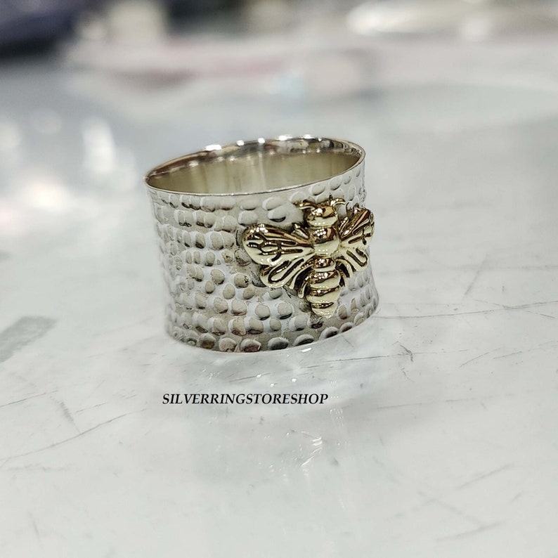 May include: A silver ring with a textured finish and a gold bee design. The ring is wide and has a hammered look.