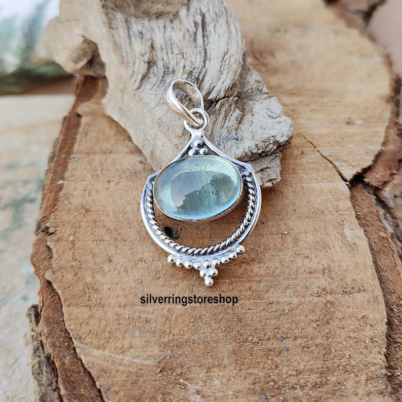 May include: A silver pendant with a light blue oval gemstone set in a decorative silver frame. The pendant has a bail for attaching to a chain.