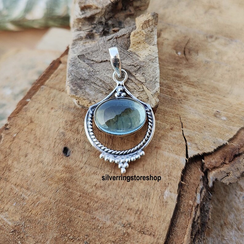 May include: A silver pendant with a light blue oval gemstone set in a decorative silver frame. The pendant is on a wooden surface.