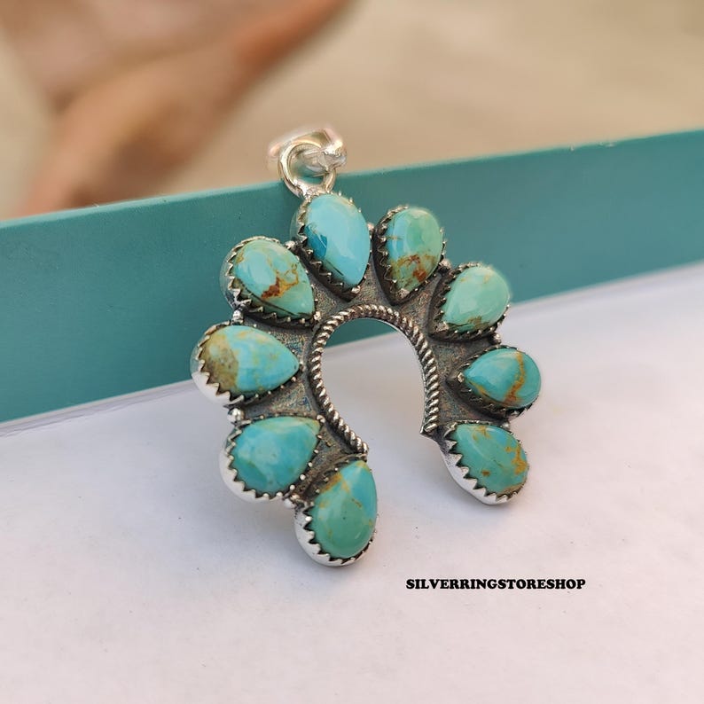 Pu&ograve; includere: Un ciondolo argentato a forma di ferro di cavallo, ornato con pietre turchesi a forma di goccia. Il ciondolo presenta un design dettagliato e strutturato e un anello per una catena. Il testo "SILVERRINGSTORESHOP" &egrave; visibile in basso.
