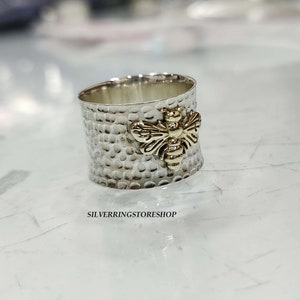 May include: A silver ring with a textured finish and a gold bee design. The ring is wide and has a hammered look.