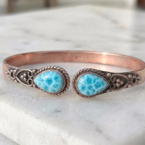 May include: Open cuff bracelet with a copper finish. The bracelet features two teardrop-shaped, light blue larimar stones. The ends of the bracelet have decorative metalwork with a rope-like texture around the stones. The bracelet is displayed on a white marble surface.