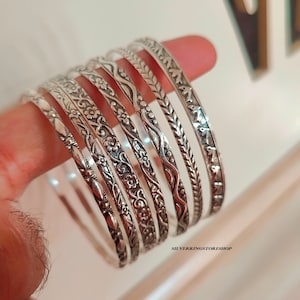 May include: Set of seven silver bangles with intricate floral and geometric designs. The bangles are stacked on top of each other and have a shiny finish.