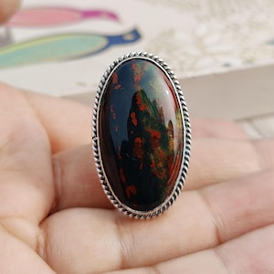 以下が含まれることがあります： 楕円形の緑と赤の宝石がついたシルバーリング。リングは装飾的なねじれたバンドをしています。