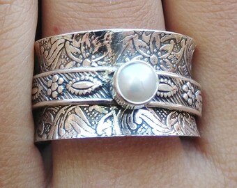 Könnte beinhalten: Ein silberner Drehring mit einer weißen Perle in der Mitte. Der Ring ist mit filigranen Blumendesigns verziert.