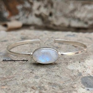 K&ouml;nnte beinhalten: Ein silbernes Armband mit einem zentralen, ovalen Mondstein, der einen durchscheinenden, irisierenden blauen Schimmer aufweist. Das Metallband ist glatt und poliert und hat eine offene R&uuml;ckseite. Der Text silverringstoreshop ist links sichtbar.