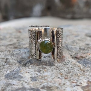 May include: A silver ring with a green gemstone set in a wide band. The band is decorated with an intricate floral pattern.