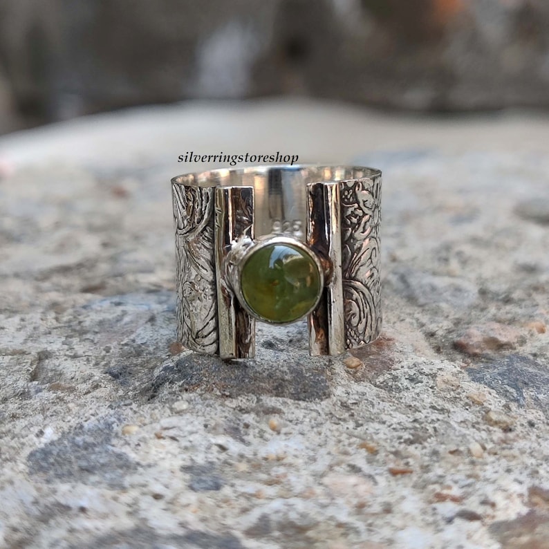 May include: A silver ring with a green gemstone set in a wide band. The band is decorated with an intricate floral pattern.